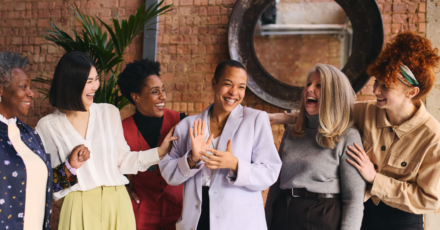 Diverse group of six women laughing together in front of an exposed brick wall with a round mirror