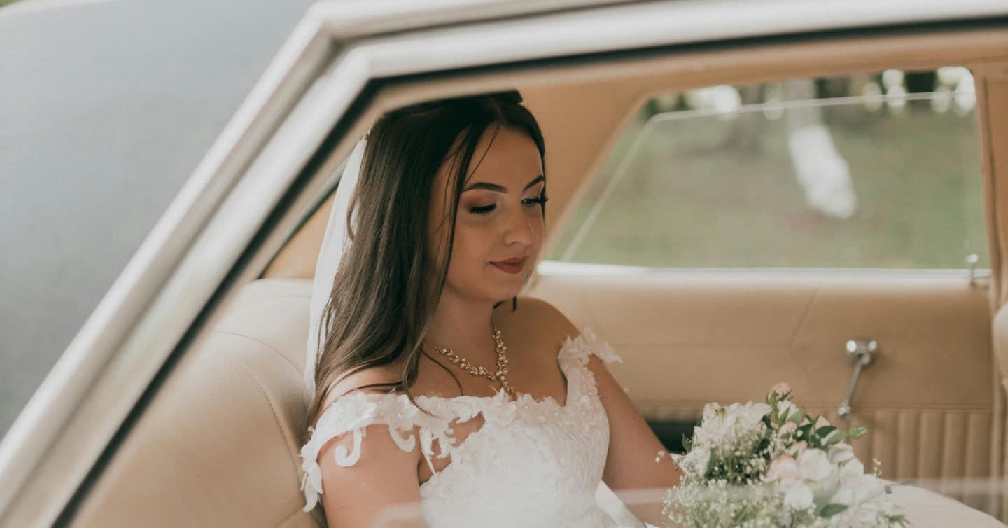 Bride with long dark hair in a white lace wedding dress holding a bouquet while seated in a vintage car
