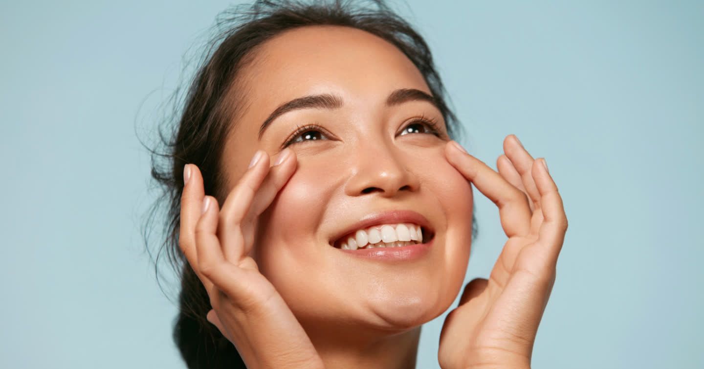 Smiling woman with glowing skin gently touching her cheeks against a soft blue background