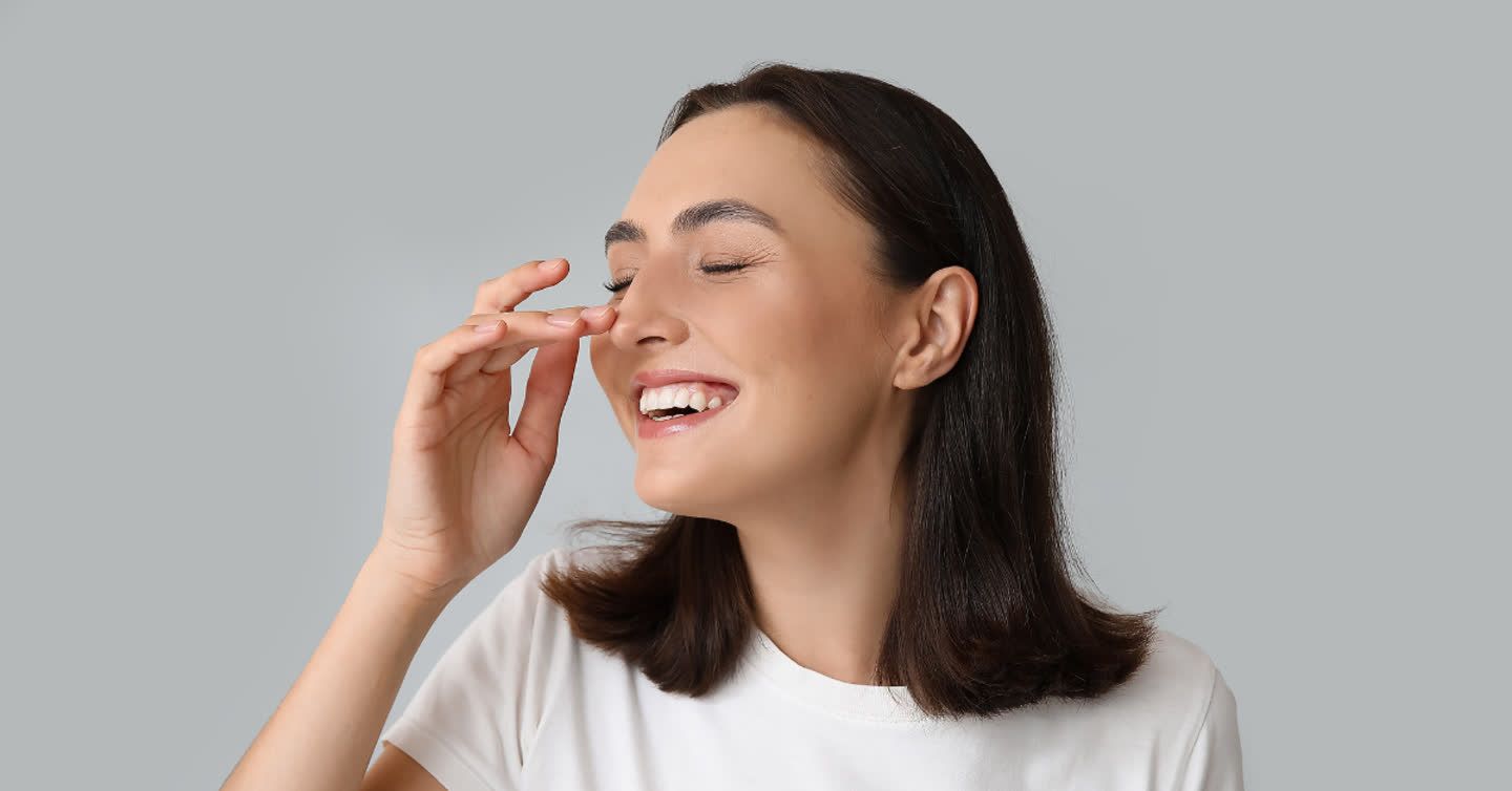 Smiling woman gently touching her nose after non-surgical rhinoplasty treatment against a grey background