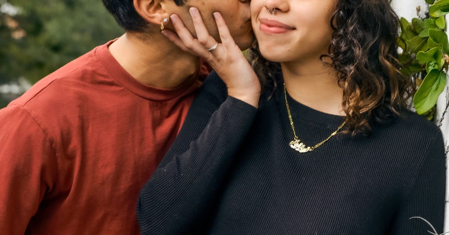 Smiling woman with curly hair and gold necklace gently touching a man's face as he kisses her cheek outdoors