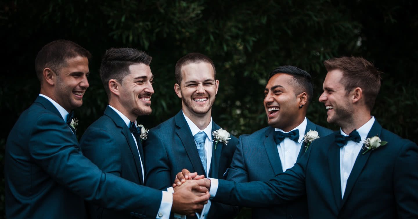 Groom and four groomsmen in navy suits with boutonnieres laughing together outdoors at a wedding