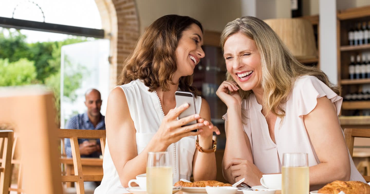 Two women laughing together at a café table with croissants and drinks while looking at a smartphone