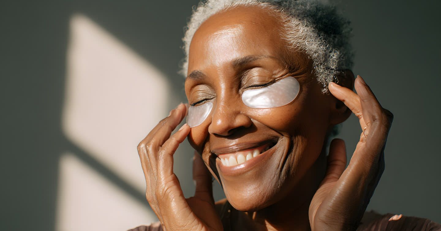 Smiling mature woman with grey hair applying under-eye patches in natural sunlight