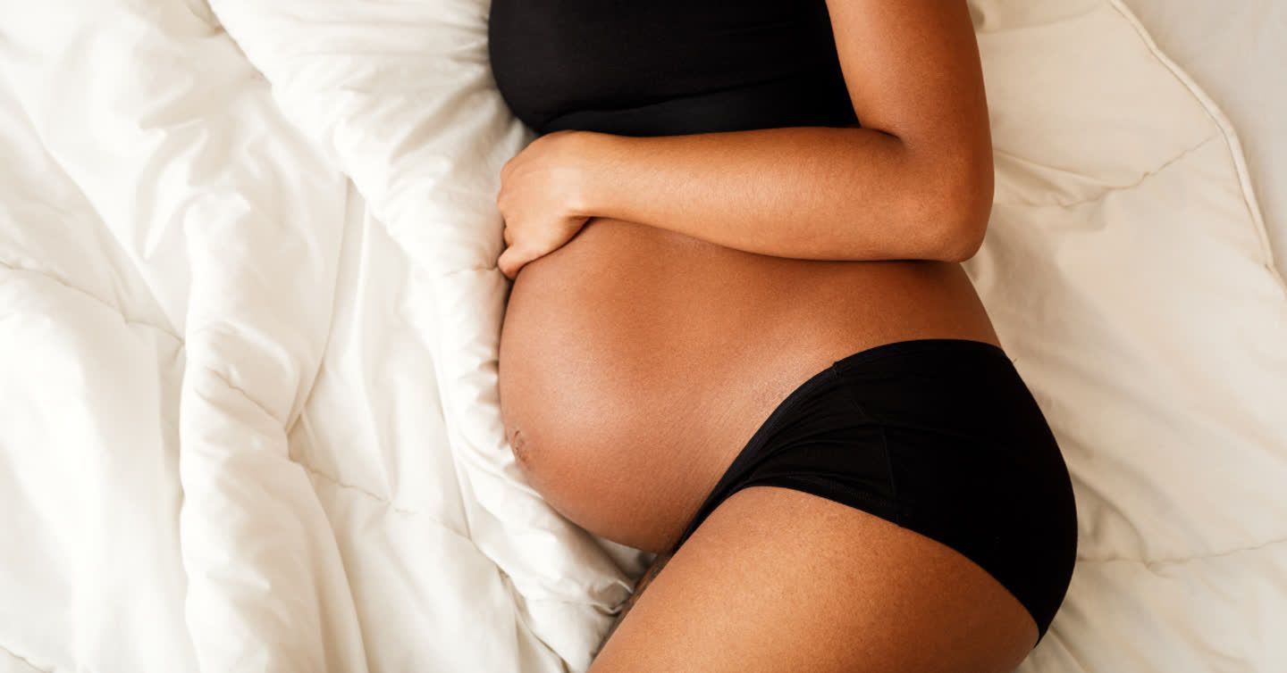 Pregnant woman lying on white bedding cradling her bare bump, showing smooth skin on her growing belly