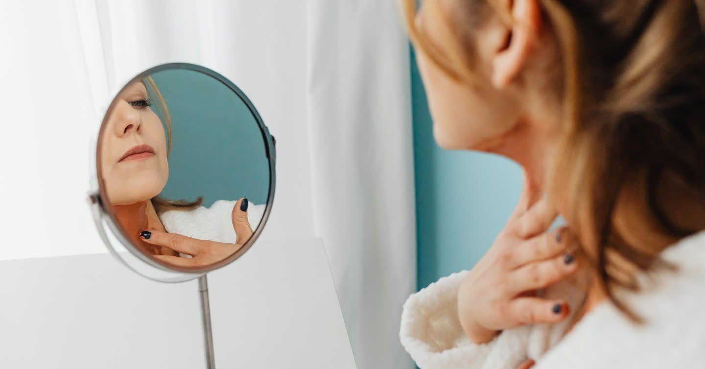 Woman in white robe examining her neck and jawline closely in a round vanity mirror