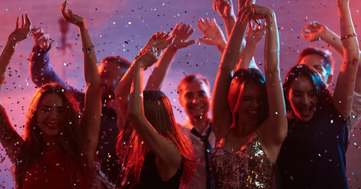 Group of friends dancing with arms raised under colourful lights as confetti falls around them at a nightclub