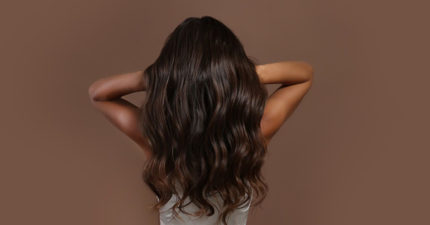 Back view of a woman with long, shiny, wavy dark brown hair against a warm brown background