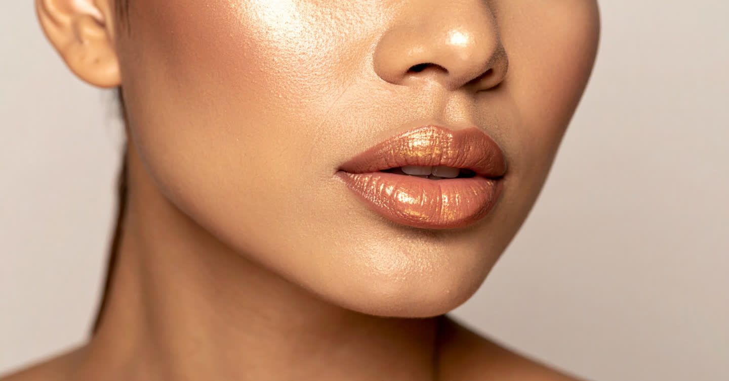 Close-up of a woman's lower face showing a smooth, defined jawline with glowing skin and gold-shimmer lip gloss