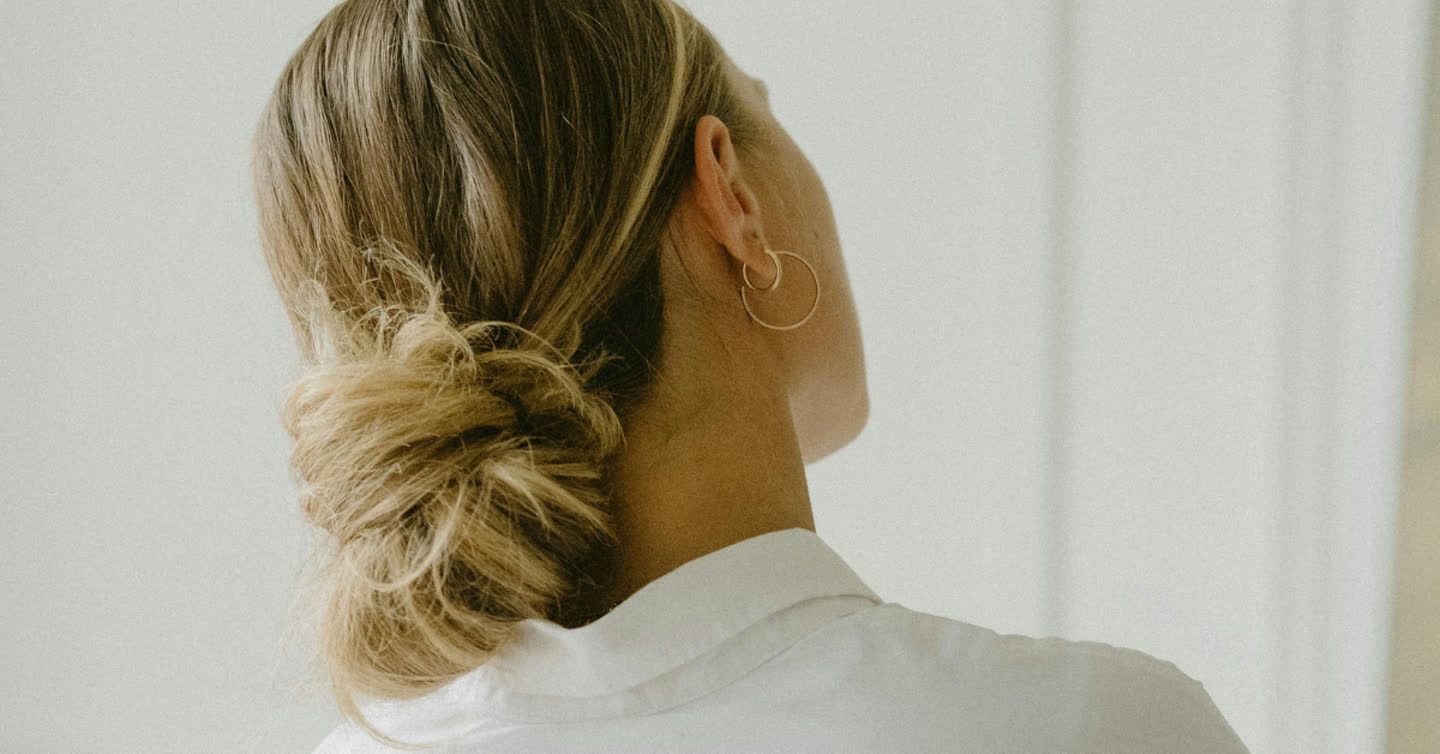 Woman seen from behind with blonde hair in a low bun, wearing a gold hoop earring and white shirt