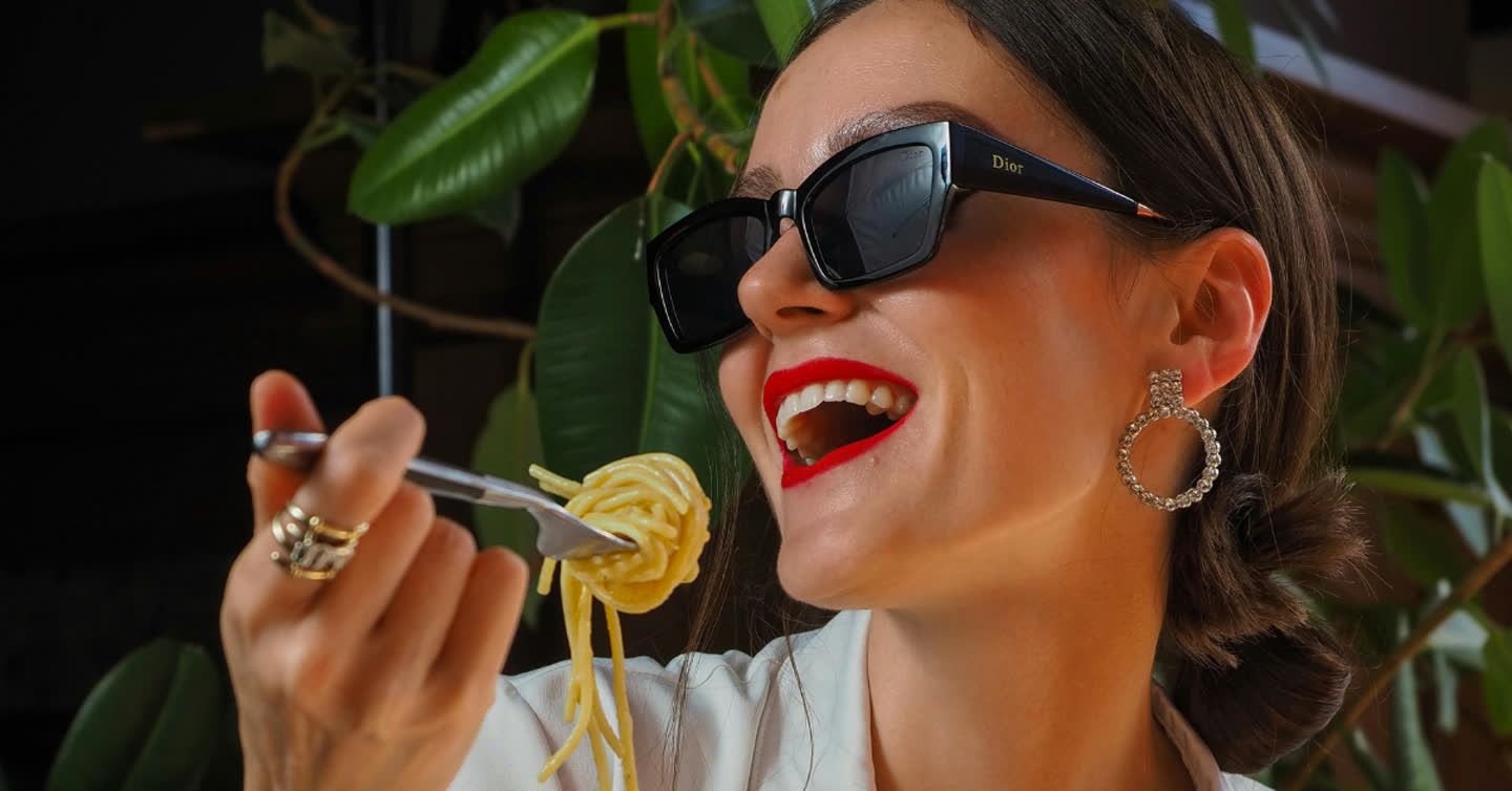 Smiling woman in Dior sunglasses and red lipstick twirling pasta on a fork with green foliage behind her
