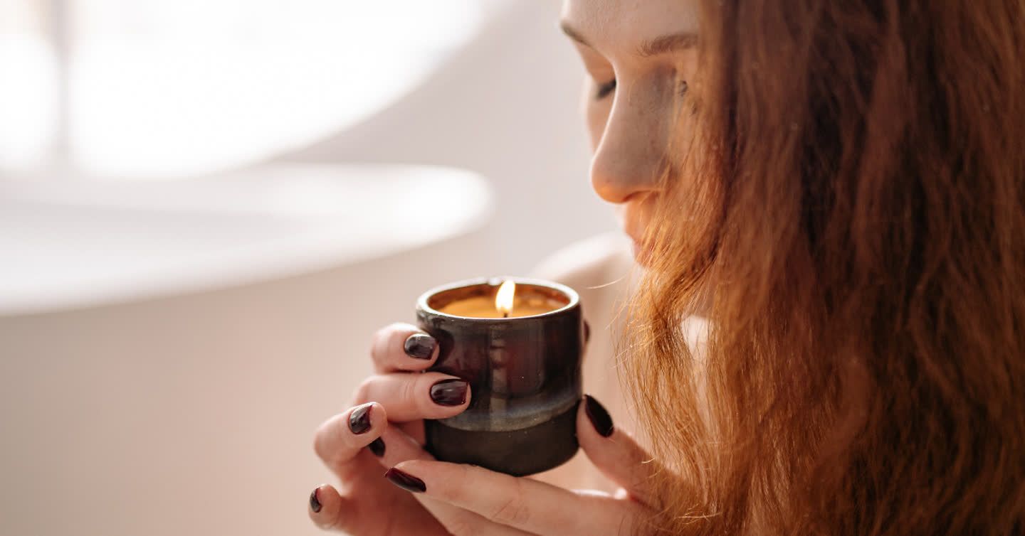 Woman with red hair holding a lit scented candle in a dark ceramic jar close to her nose, smelling the fragrance