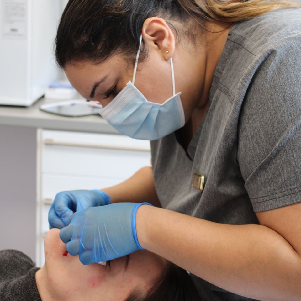 Practitioner in grey scrubs and blue gloves performing a facial treatment on a patient lying on a clinic bed