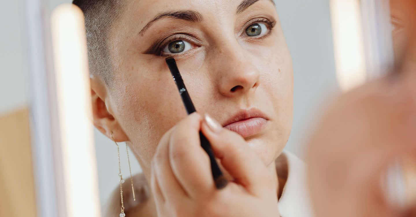 Woman with short hair applying eyeliner pencil beneath her eye while looking into a mirror with warm lighting