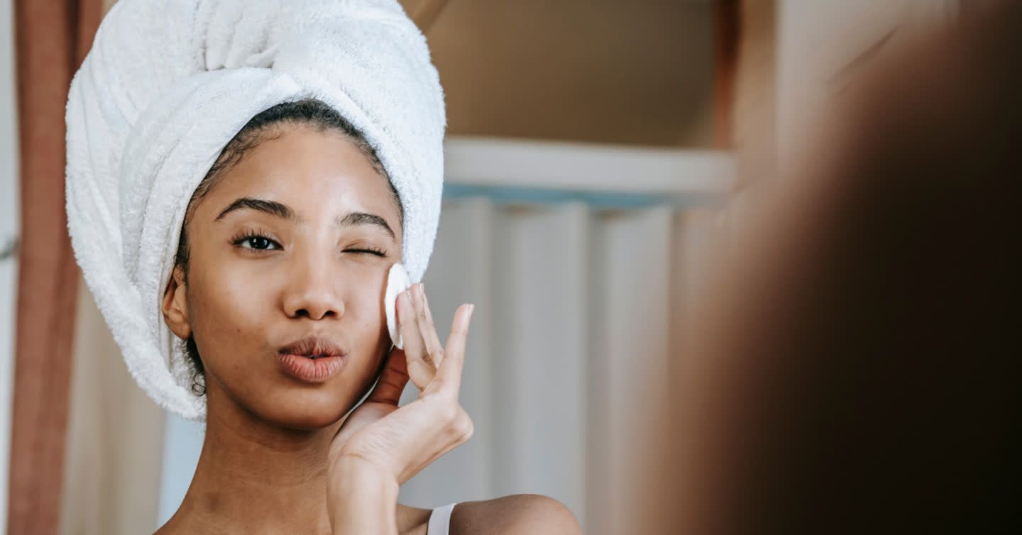 Woman with towel-wrapped hair applying skincare product to her cheek while looking in a bathroom mirror