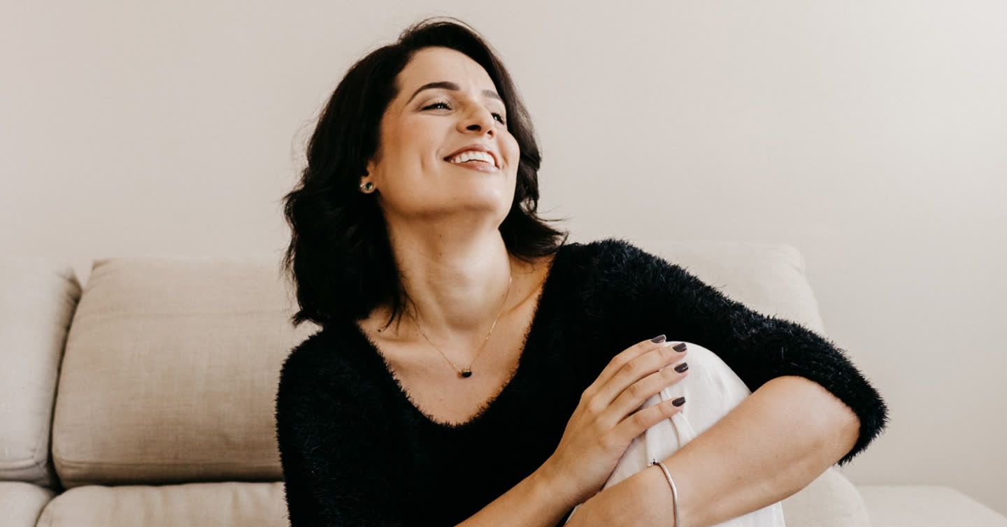 Smiling woman with dark hair sitting on a cream sofa, wearing a black jumper and delicate necklace, showing a smooth neck