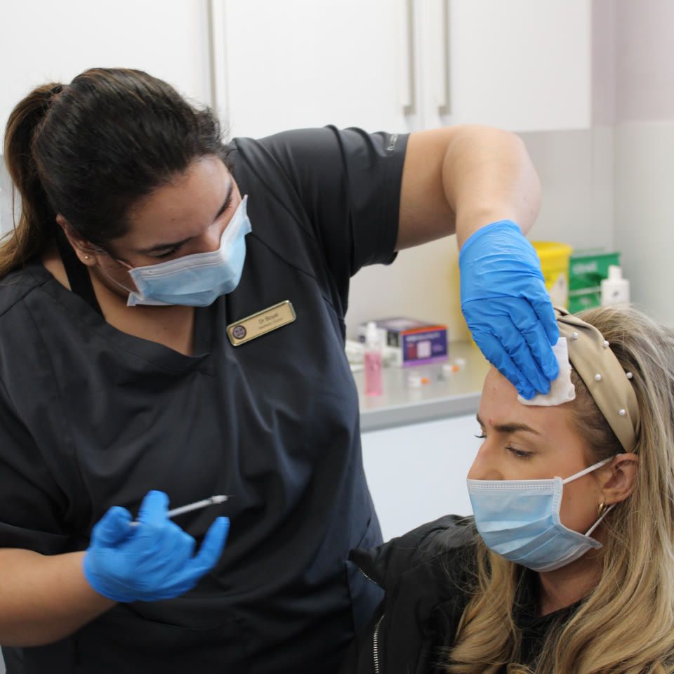 Practitioner in scrubs applying a gauze pad to a patient's forehead before fat dissolving injections in a clinical treatment room
