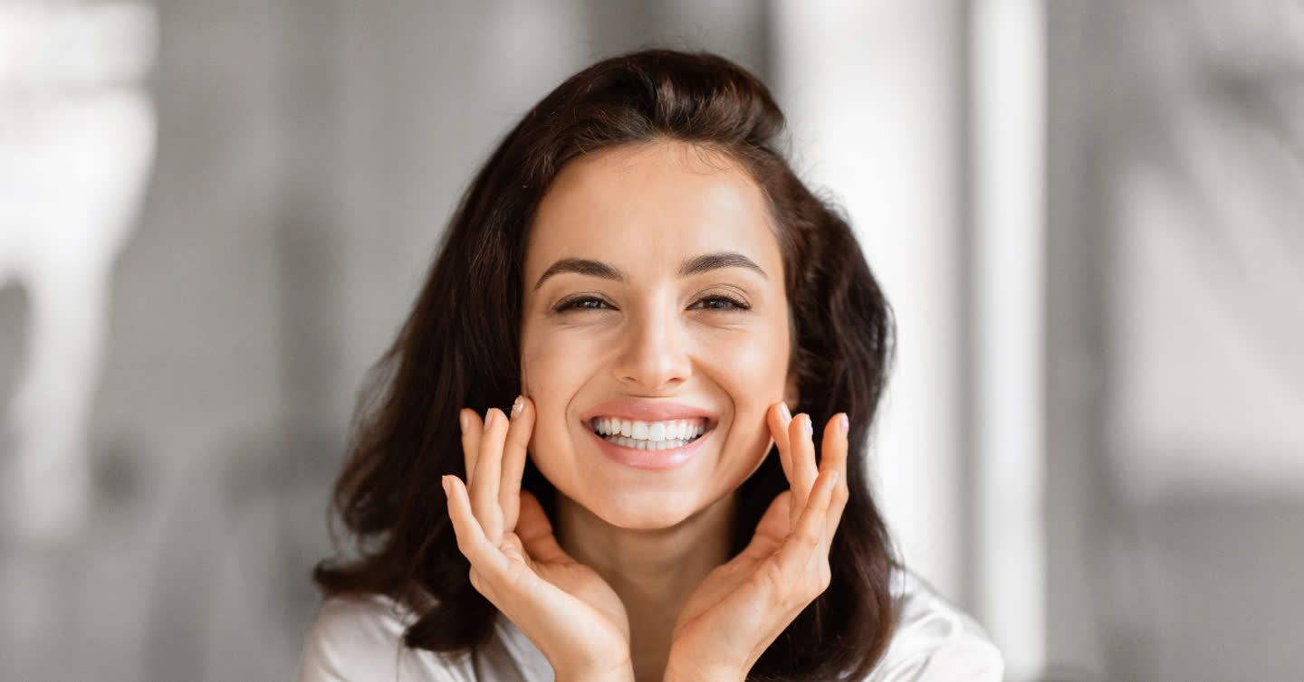 Smiling woman with dark hair gently touching her face with both hands, showcasing smooth radiant skin after facial fat transfer
