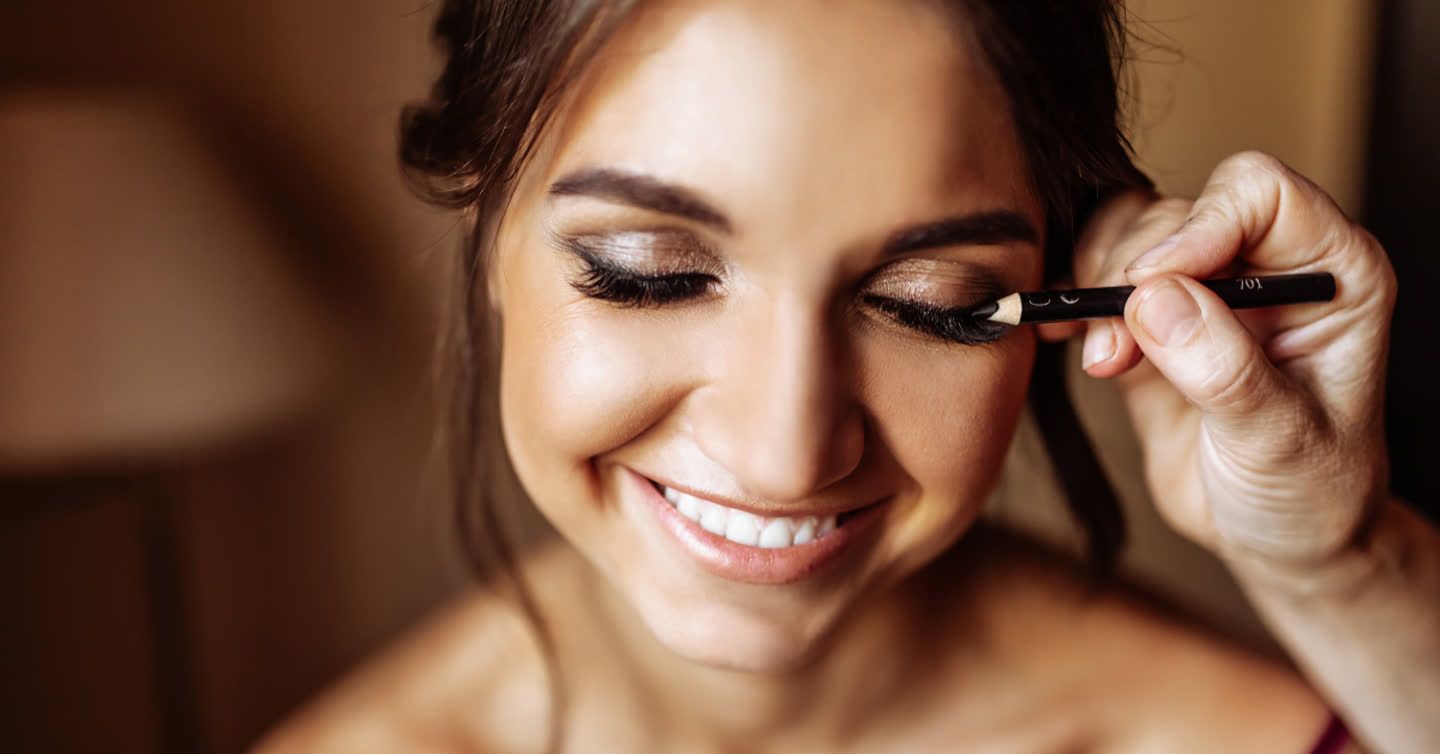 Smiling woman with eyes closed as a makeup artist applies eyeliner pencil to her lower lash line