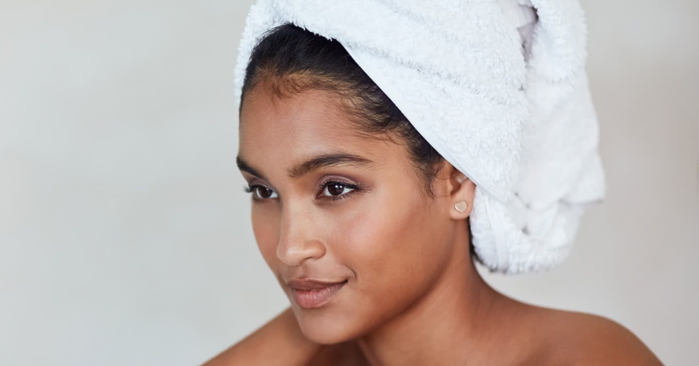 Woman with glowing skin and a white towel wrapped around her hair, looking over her shoulder with a soft smile
