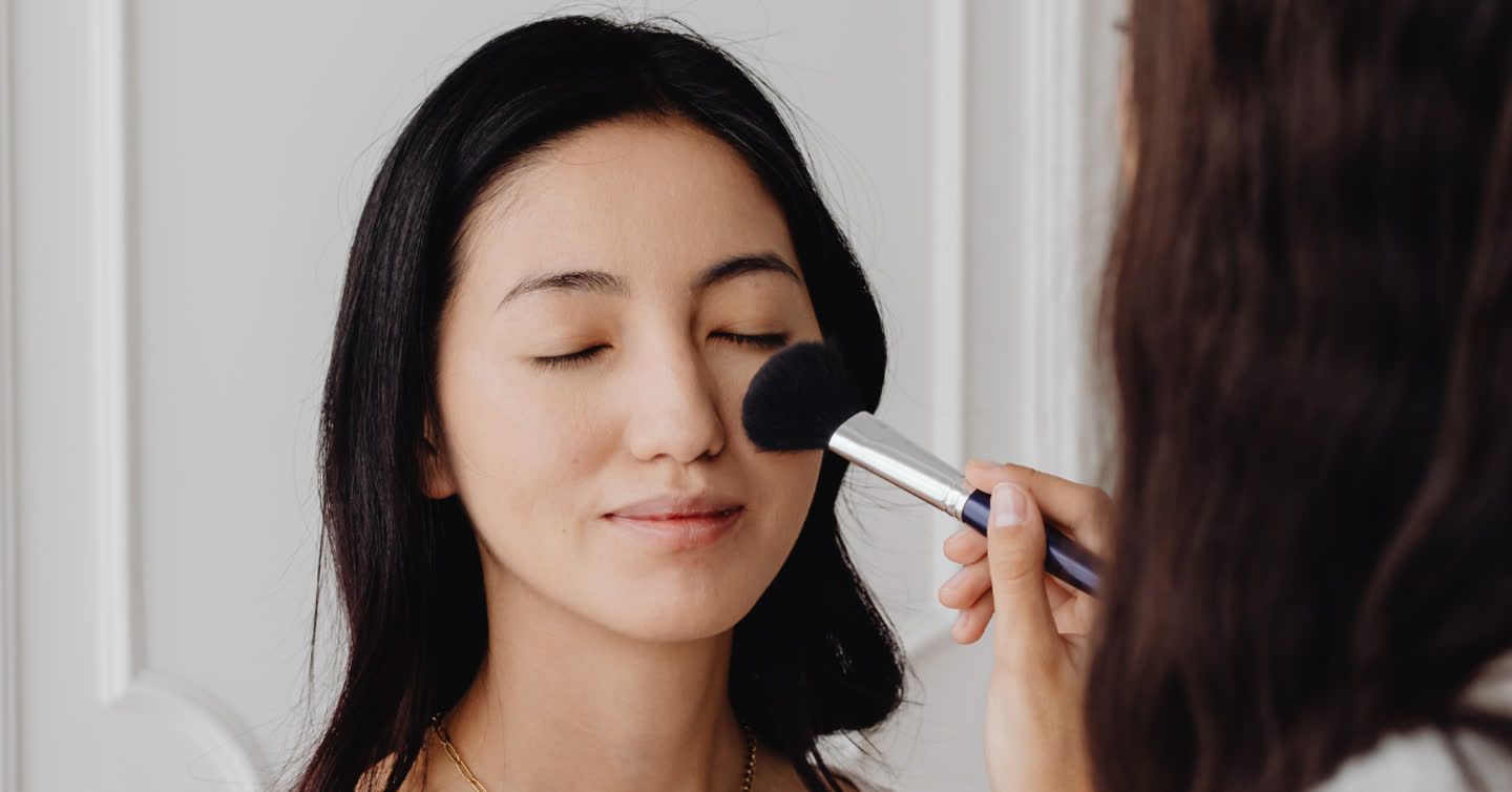 Makeup artist applying powder with a large brush to a woman's cheek as she closes her eyes