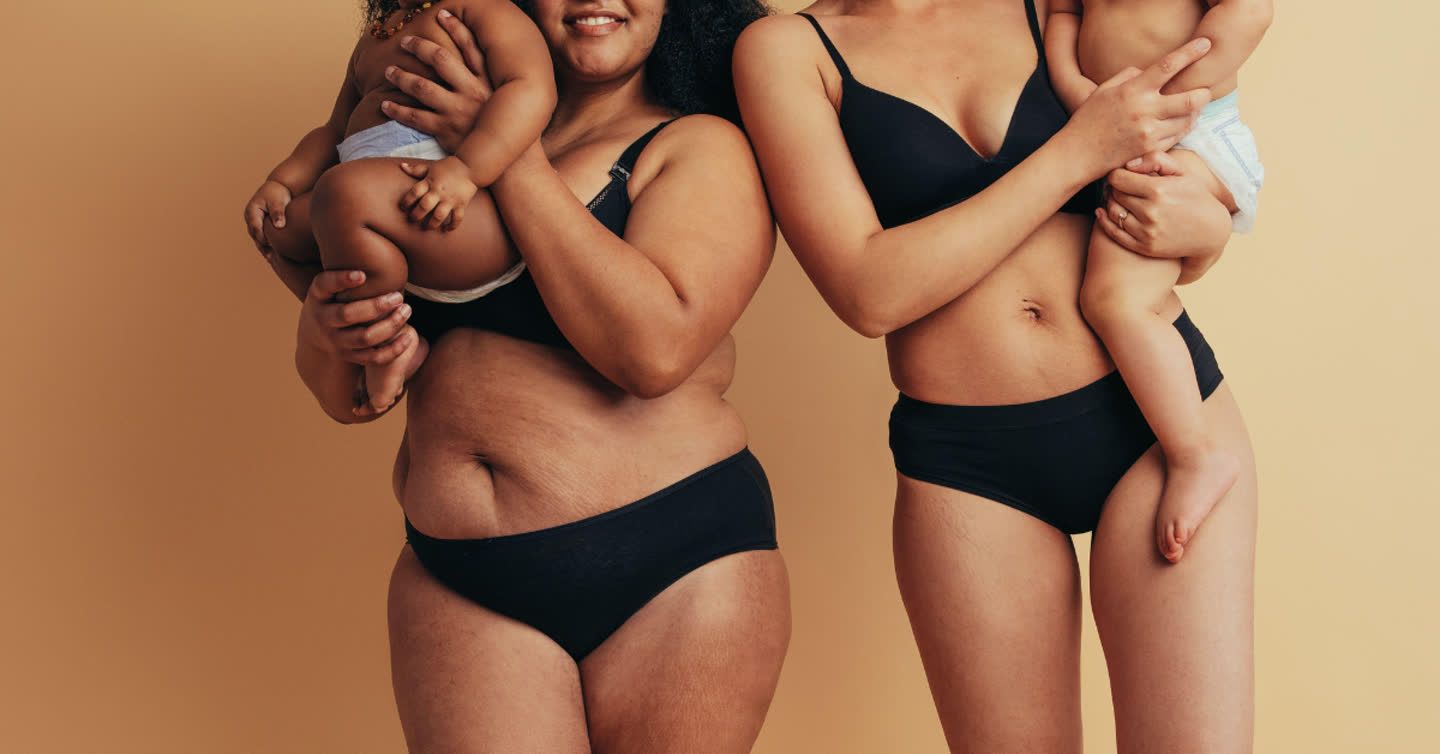 Two mothers with different postpartum body types wearing black underwear, each holding a baby against a warm beige background