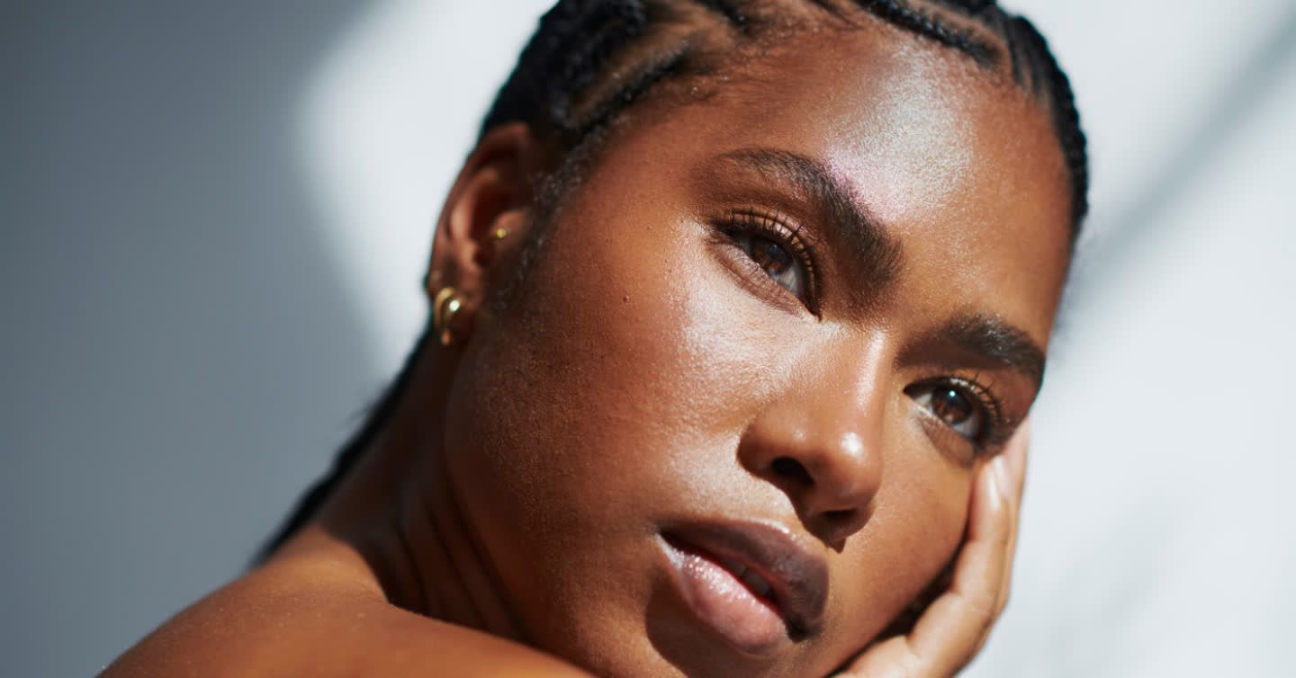 Close-up of a woman with braided hair and glowing skin resting her chin on her hand in natural sunlight