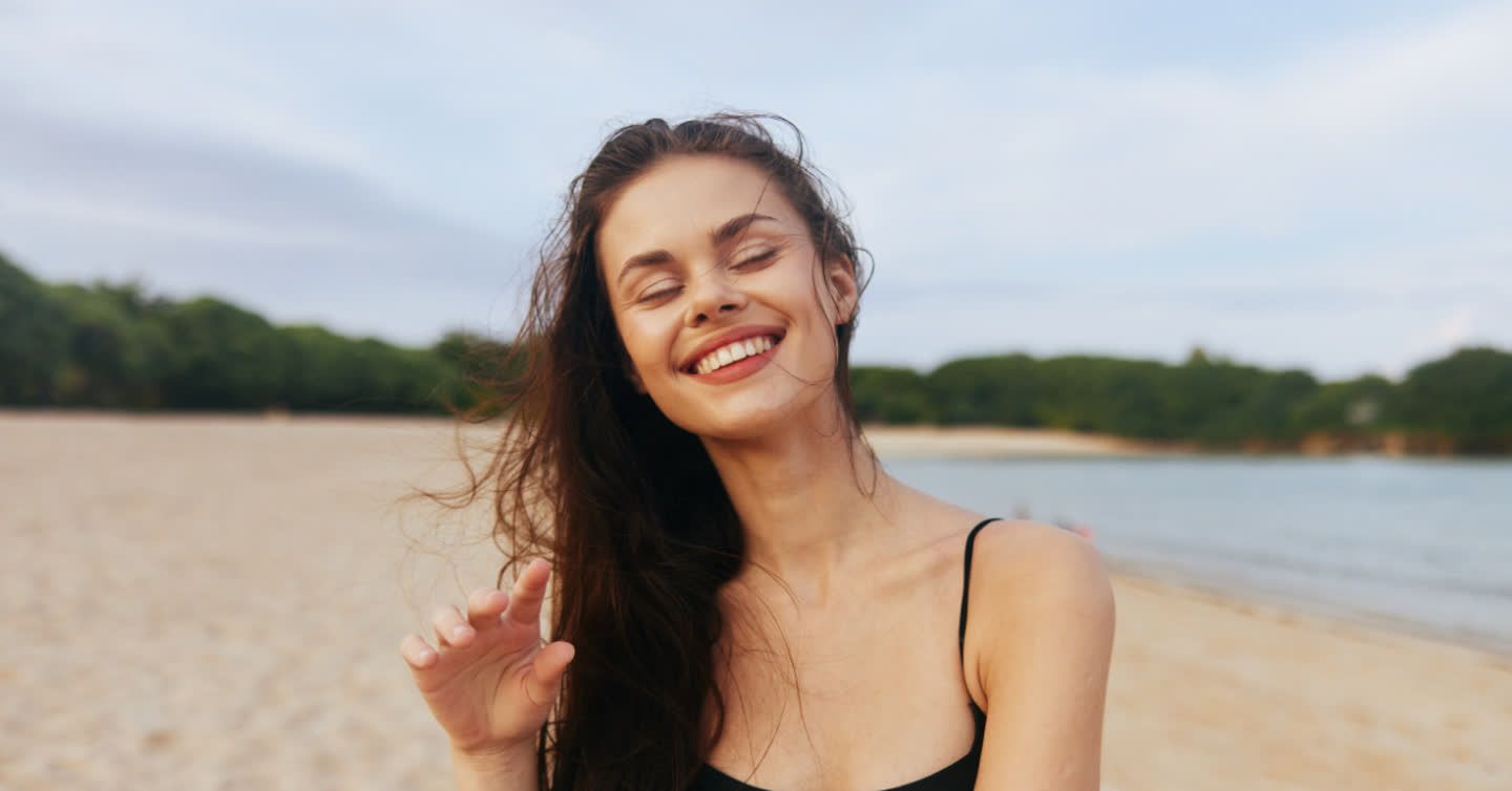 Smiling woman with long dark hair blowing in the wind on a sandy beach with eyes closed and a defined jawline