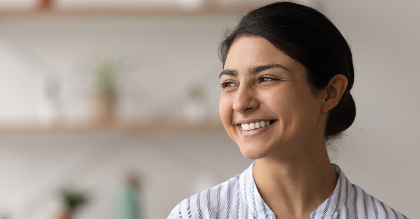 Smiling young woman with dark hair tied back, looking to the side in a bright room with shelves