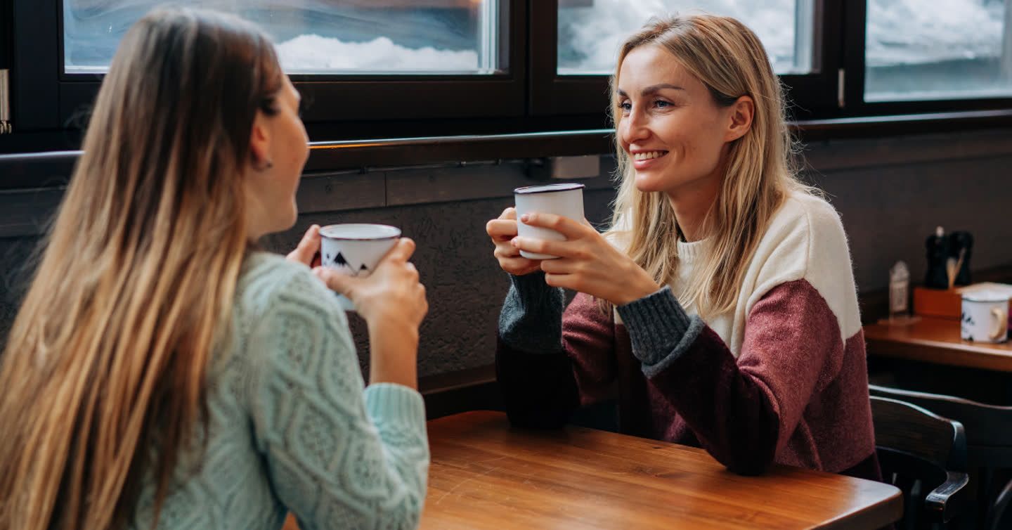 Two smiling women in cosy jumpers chatting over hot drinks at a wooden café table by a window