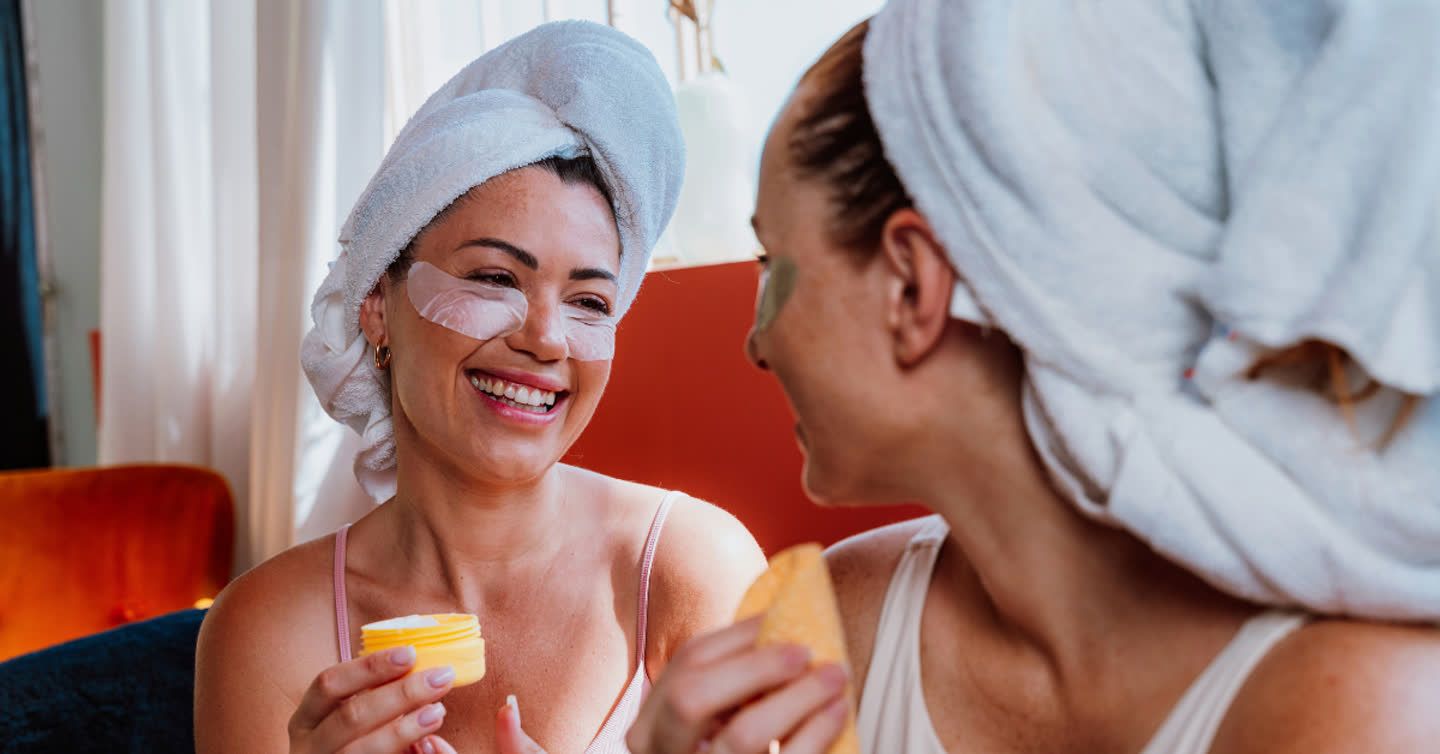 Two smiling women with towel-wrapped hair wearing under-eye patches and holding skincare products during an at-home beauty treatment