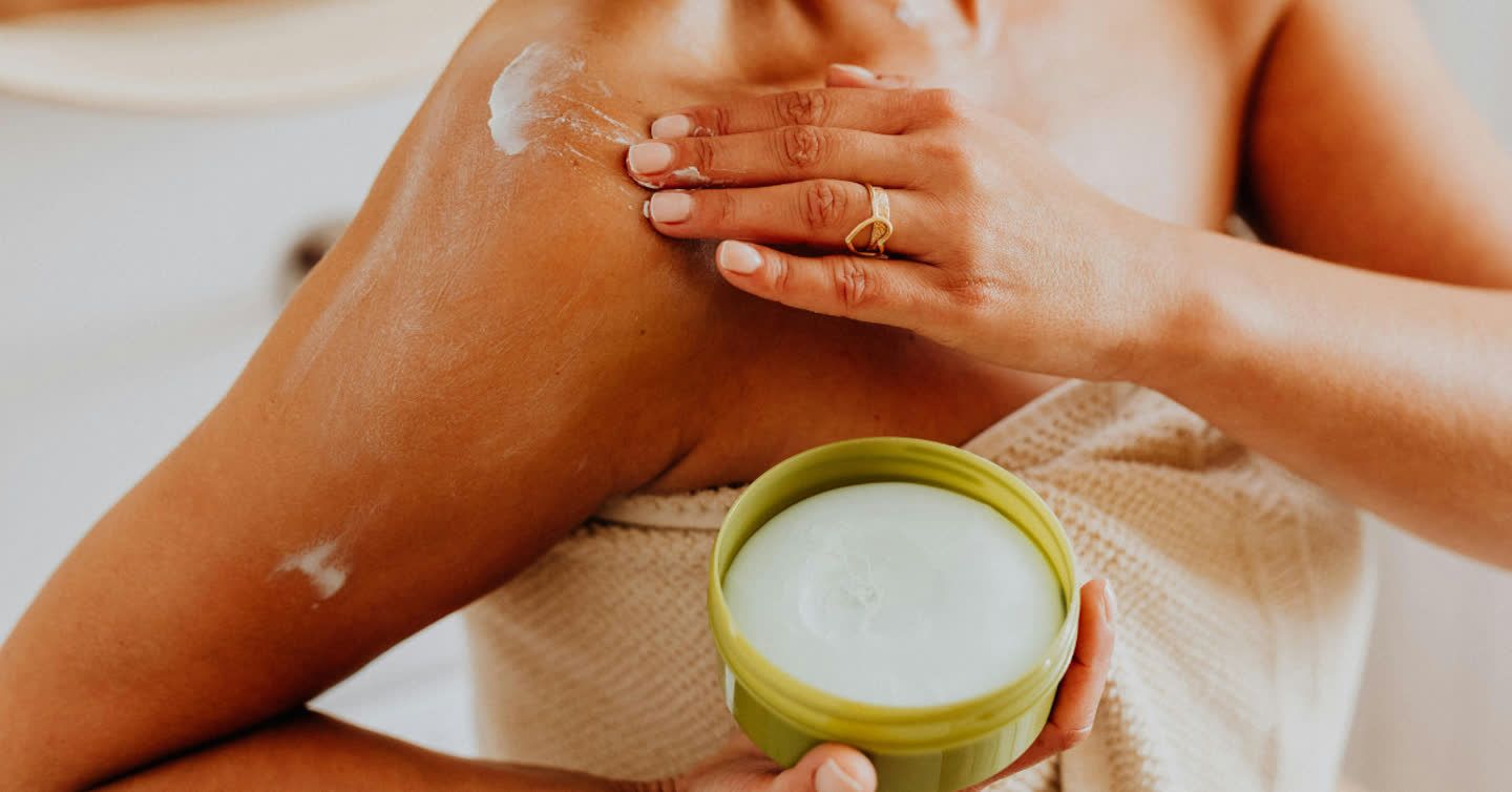 Woman applying white moisturising cream from a green jar onto her shoulder while wrapped in a towel