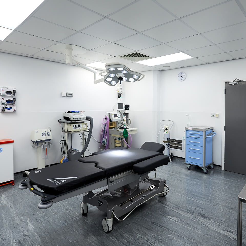 Modern surgical theatre with operating table, overhead LED light, anaesthesia equipment, blue supply trolley and clinical waste bins