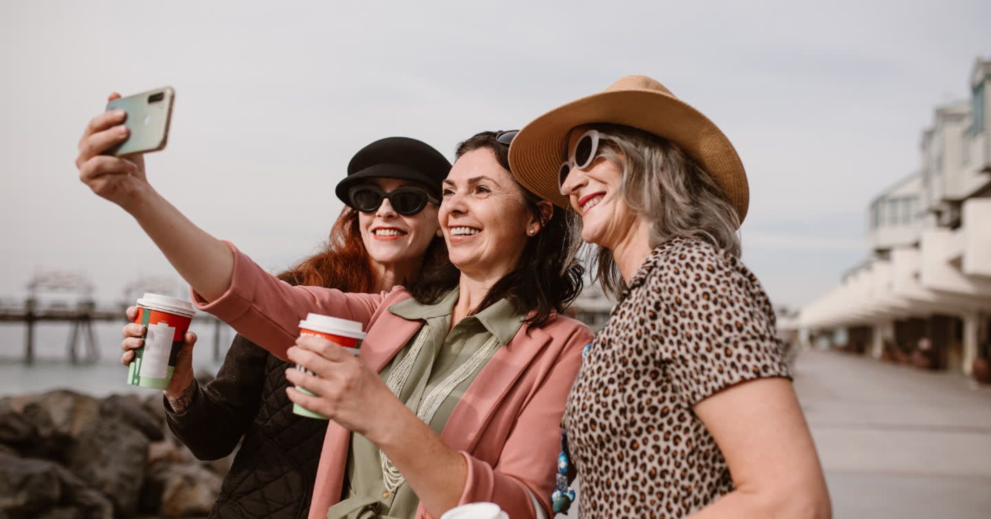Three smiling women in sunglasses and hats taking a selfie while holding coffee cups on a seaside promenade