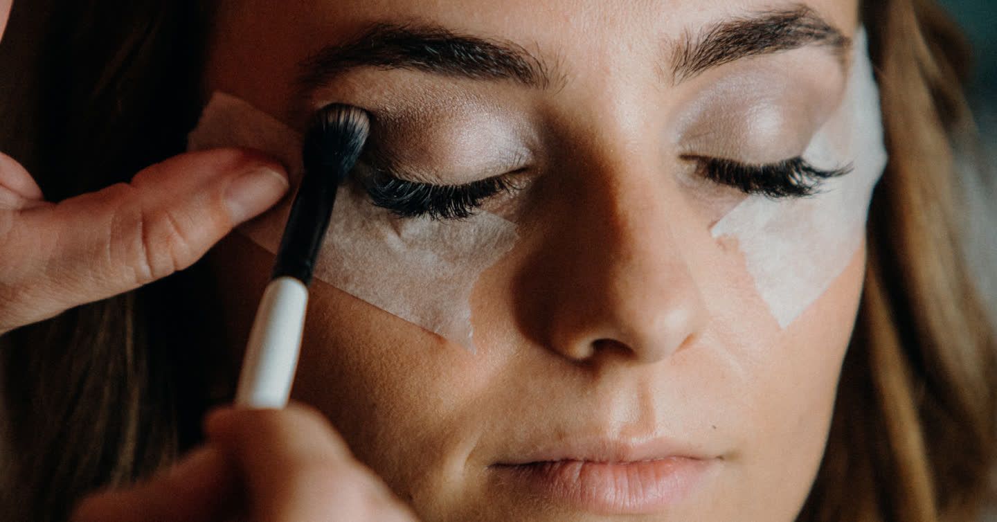 Makeup artist applying eyeshadow with a brush to a woman's closed eyelid with tape guides on her cheeks