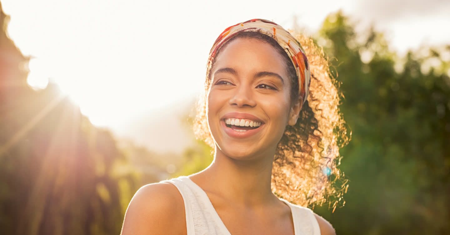 Young woman with curly hair and headband smiling outdoors in golden sunlight with green trees behind