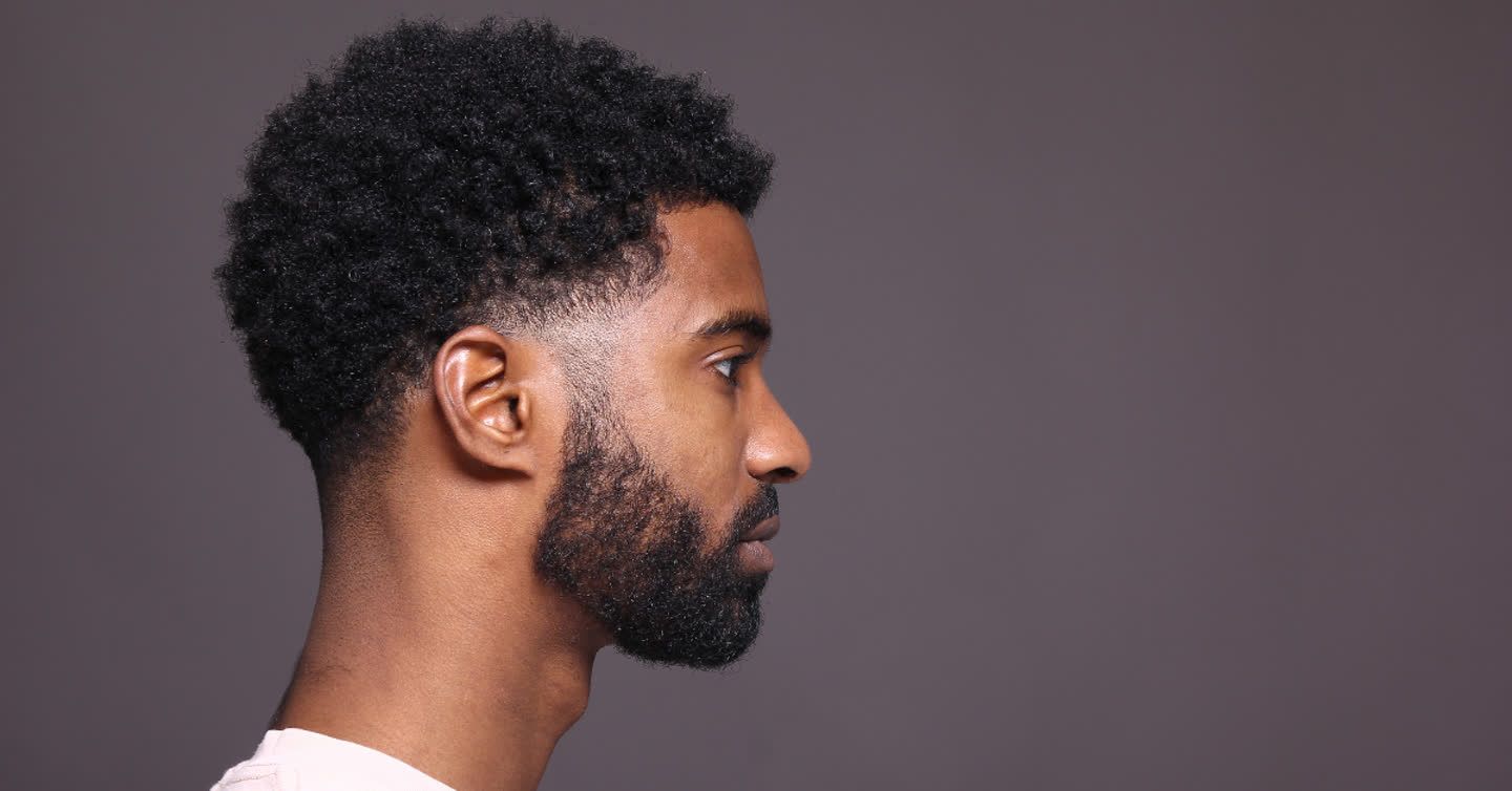 Side profile of a young man with a natural afro hairstyle and beard against a grey background, showing his ear