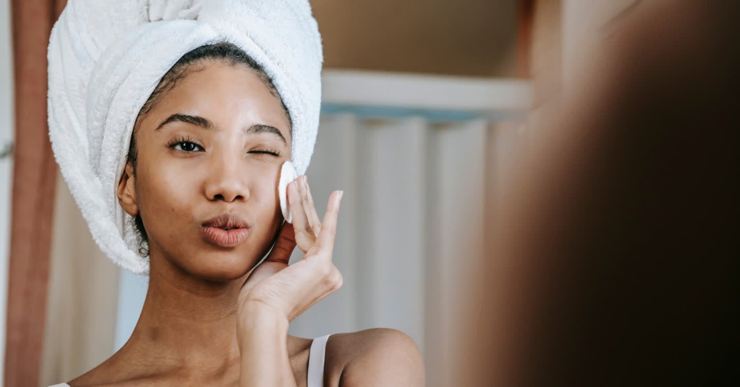 Woman with towel-wrapped hair applying a cotton pad to her cheek while looking in a bathroom mirror