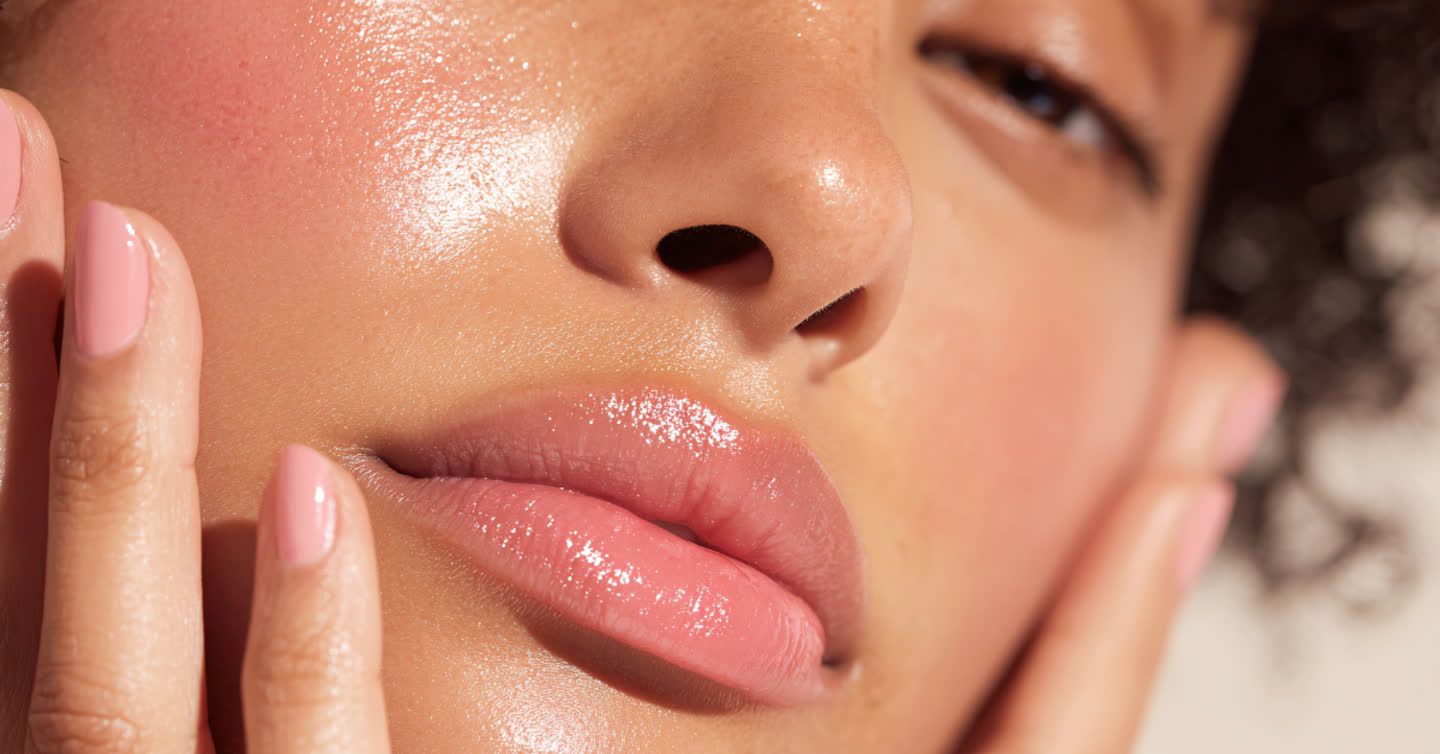 Close-up of a woman with glowing, hydrated skin and glossy pink lips gently touching her cheek with manicured fingers