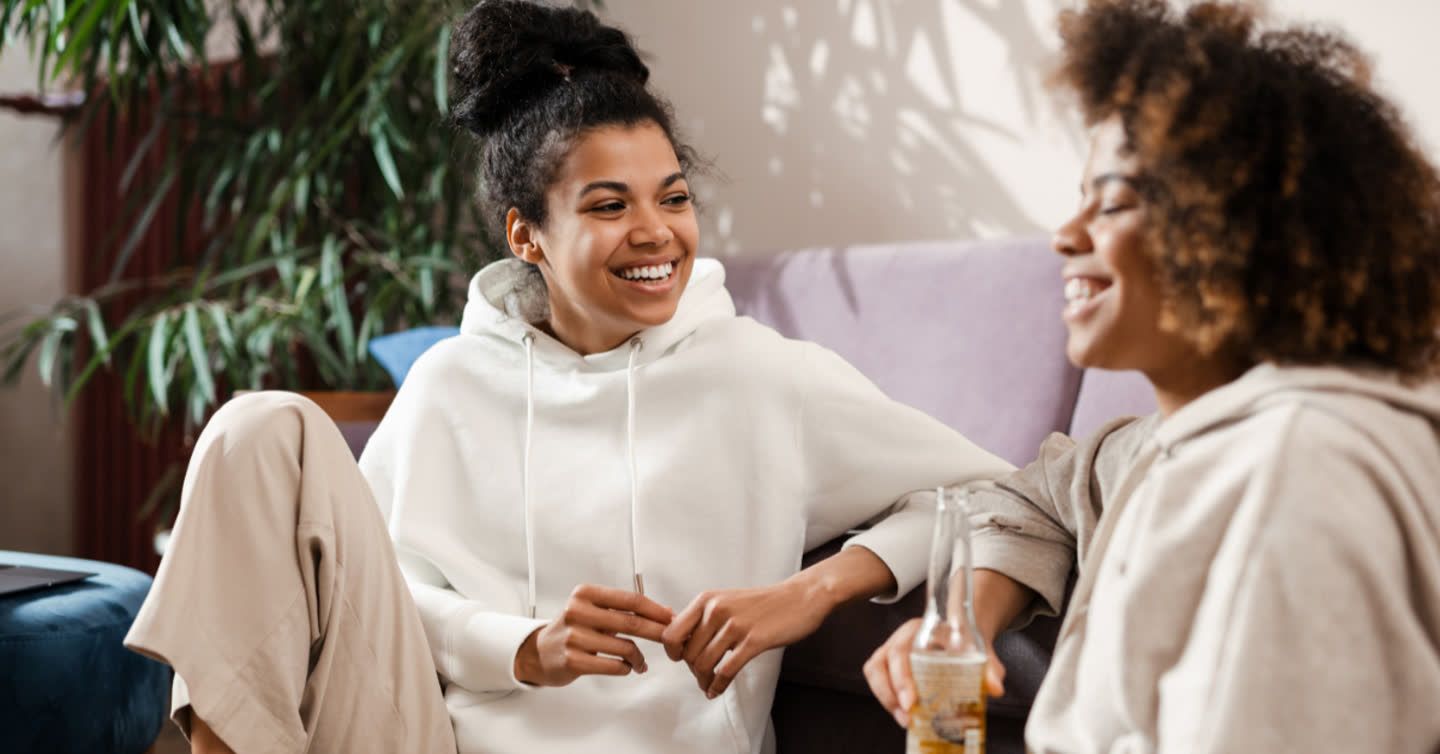 Two young women laughing together on a sofa, one wearing a white hoodie and the other holding a glass bottle