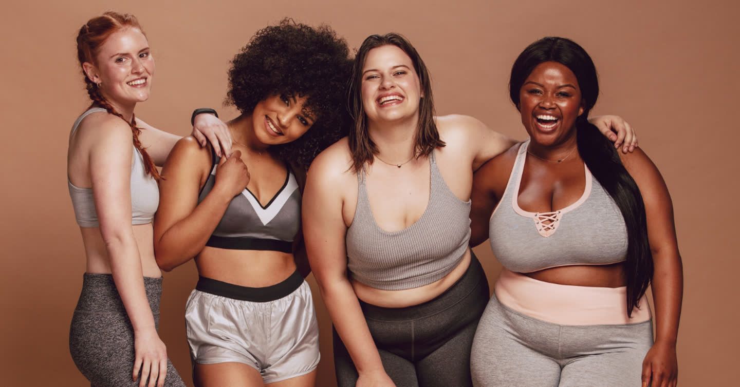 Four diverse women with different body types laughing together in sports bras and activewear against a warm brown backdrop