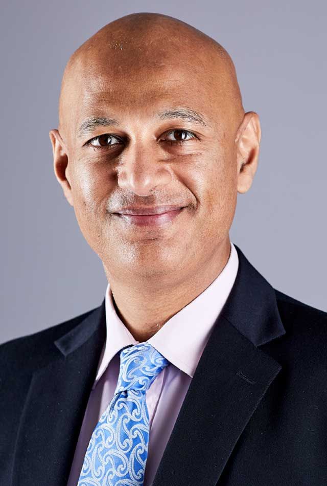 Professional headshot of a smiling man in a dark suit, light pink shirt, and blue patterned tie