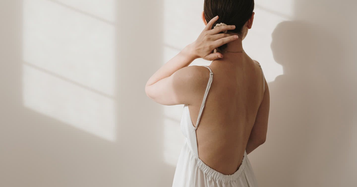 Woman seen from behind in a low-back white dress touching her neck in a sunlit room