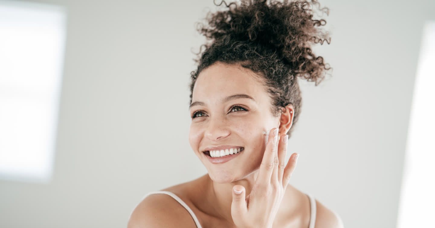 Smiling young woman with curly hair touching her cheek while applying moisturiser in a bright room