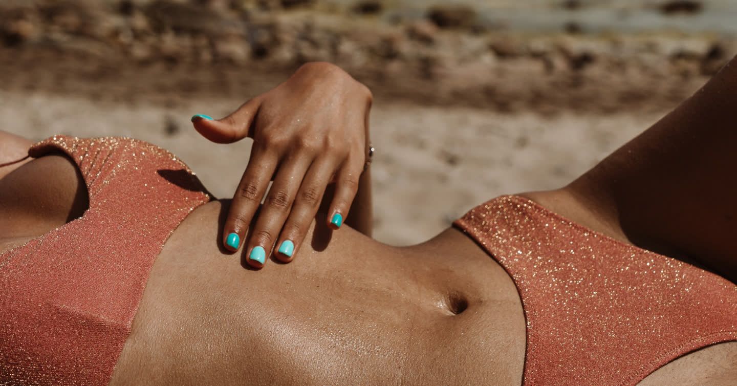 Woman's hand with turquoise nails resting on her toned stomach while sunbathing in a sparkly coral bikini