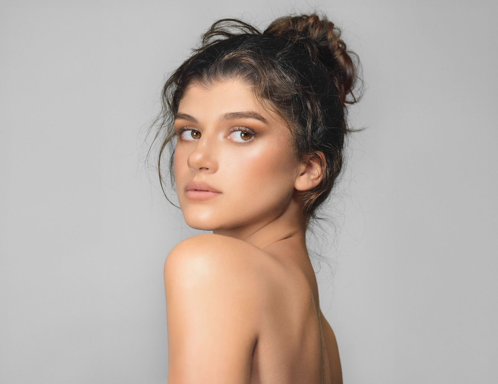 Young woman with messy bun hairstyle looking over her bare shoulder, showcasing clear glowing skin