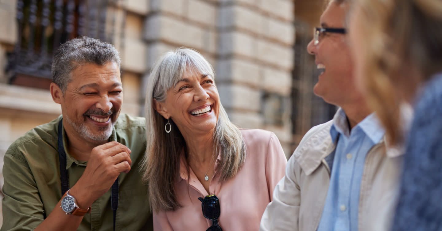 Group of middle-aged friends laughing together outdoors, woman with grey hair centre, in front of a sandstone building