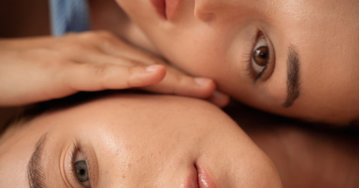 Close-up of two women with smooth, clear skin resting their faces together, one with brown eyes and one with blue eyes