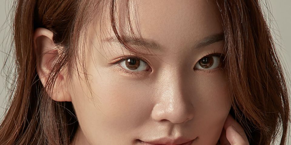 A woman with glowing skin and long auburn hair smiling softly, resting her chin on her hand against a neutral grey background