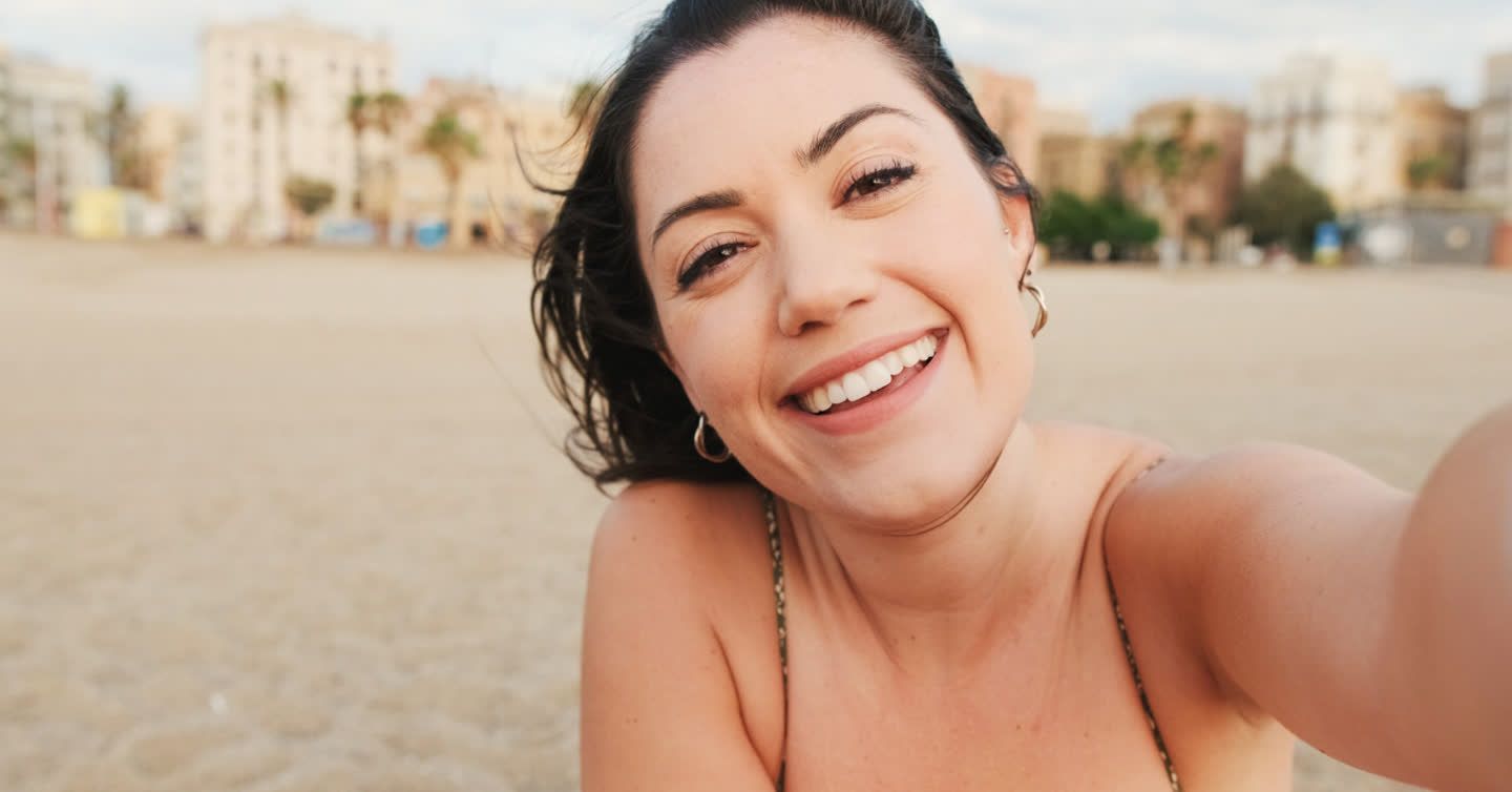 Smiling woman taking a selfie on a sandy beach with buildings and palm trees in the background